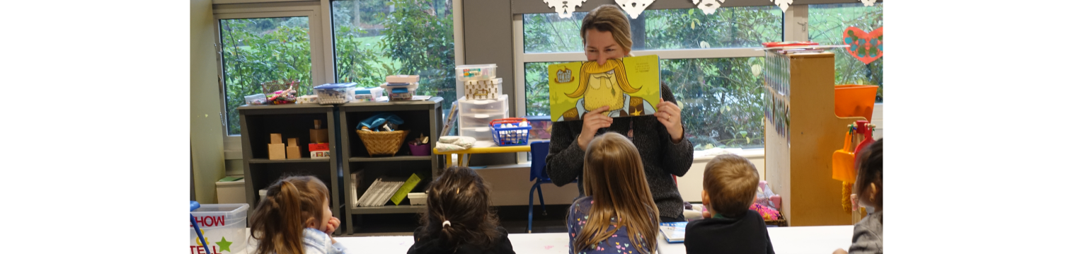 a teacher holding a book to their face with an illustration of a mustache, reading to a group of preschool children