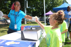 a counselor in a blue shirt with pink hair, smiling as she hands an ice cream cone to a child wearing a neon yellow shirt